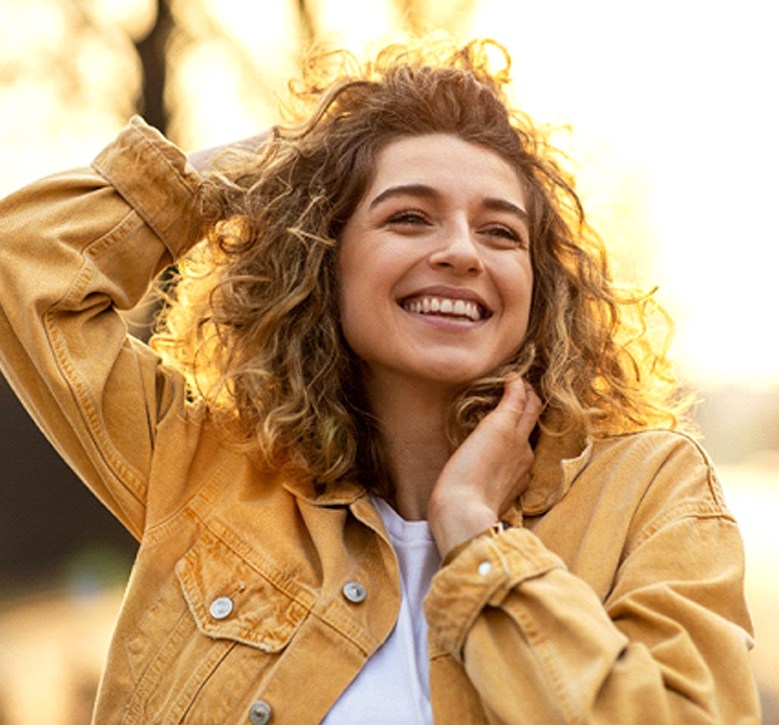 A young female wearing a mustard colored jacket and standing outside while smiling after receiving metal-free crowns in Alamo Ranch, TX