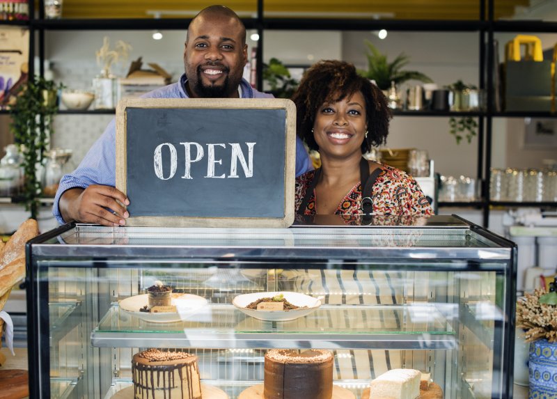 a couple standing behind a bakery counter holding a sign that reads “OPEN” 