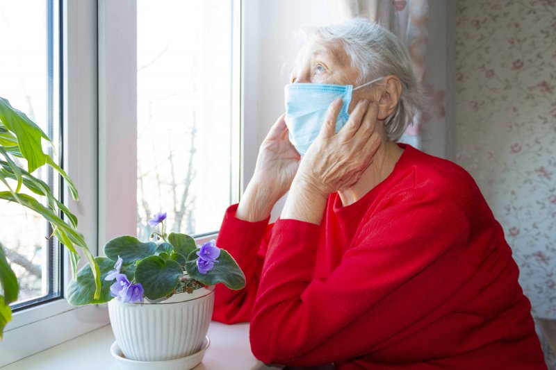 an elderly woman wearing a red sweater and staring out the window with her mask on